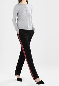 Kvinna klädd i en grå knappad kofta, svarta byxor med röda och vita sidostripes, och svarta högklackade skor, går med händerna i fickorna.