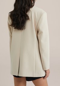 Beige oversized kavaj med skräddarsydd passform, featuring axelvaddar, en ventilerad rygg och slät textur. Stylad över svarta shorts.