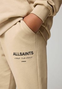 Beżowe dresy o gładkiej teksturze, z czarnym nadrukiem logo "ALLSAINTS" oraz japońskim tekstem na bocznej kieszeni.