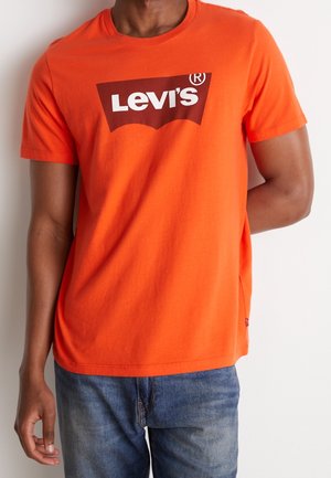 Persona vistiendo una camiseta Levi’s de color naranja brillante con jeans azules, posando contra un fondo liso de color claro.