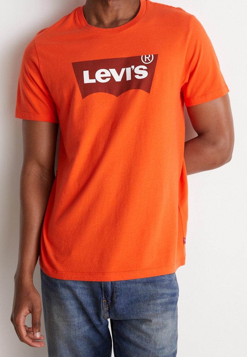 Personne portant un t-shirt Levi's orange vif avec un jean bleu, debout devant un fond uni de couleur claire.