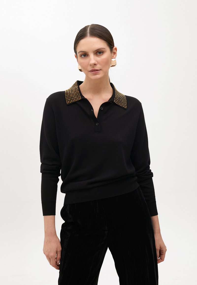 Schwarzer Pullover mit einem Knopfkragen, der mit goldenen Nieten verziert ist. Langärmlig, taillierter Schnitt, aus weichem Material, kombiniert mit schwarzen Plisséhosen.