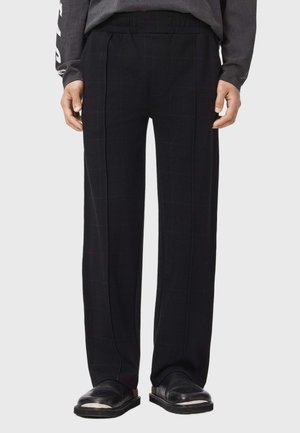 Mann trägt schwarze, gerade geschnittene Hose mit elastischem Bund und schwarze Lederschuhe ohne Schnürsenkel mit silbernen Zehenkappen.