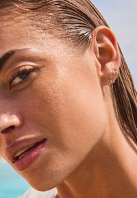 Nahaufnahme eines nassen Ohres mit drei silbernen Creolen, glatter Haut mit sichtbaren Sommersprossen und hellbraunem Haar. Der Hintergrund ist unscharf.