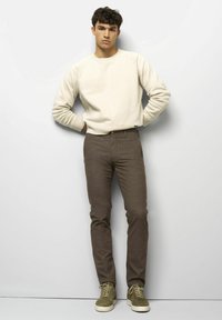 Junger Mann steht an einer weißen Wand und trägt einen cremefarbenen Pullover, braune Slim-Fit-Hosen und olivgrüne Sneakers mit weißen Sohlen.