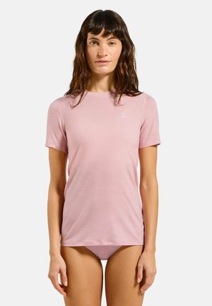 Femme aux longs cheveux bruns portant un t-shirt rose clair à manches courtes et des sous-vêtements assortis, debout devant un fond blanc uni.