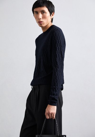 Navy gestrickter Pullover mit strukturiertem Rautenmuster, Rundhalsausschnitt und langen Ärmeln, kombiniert mit schwarzen Hosen und einer geflochtenen schwarzen Handtasche.
