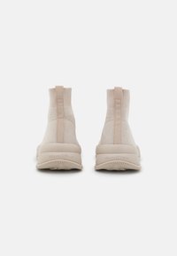 Béžové pletené kotníkové tenisky se designem připomínajícím ponožku, s poutky na zadní straně a strukturovanými gumovými podešvemi. Obsahují decentní branding na podrážce.