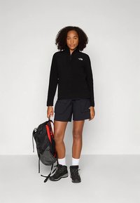 Czarna bluza z polaru z zamkiem przy kołnierzu, noszona z czarnymi szortami typu cargo. Czarne buty trekkingowe i czarno-czerwony plecak dopełniają strój.