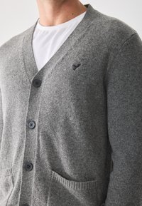 Cardigan gris hecho de tejido texturizado, con cuello en V, tres botones y dos bolsillos frontales. Detalle de logotipo sutil bordado en el pecho.