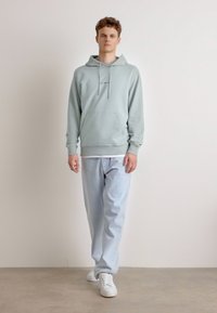 Felpa verde menta con tasca frontale, abbinata a jeans azzurri e sneakers bianche, in piedi contro uno sfondo neutro.