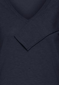 Haut à manches longues bleu marine avec un col en V, une texture côtelée et une coupe légèrement ample. Détail de couture visible sur l'épaule.