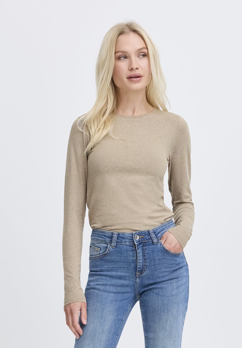 Blonde Frau, die ein beiges Langarmshirt und blaue Jeans trägt, steht mit einer Hand in der Tasche und blickt zur Seite vor einem schlichten weißen Hintergrund.