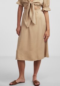 Gonna midi beige realizzata in tessuto morbido, con cintura legata e tasche laterali. Abbinata a sandali marroni con sola piatta.