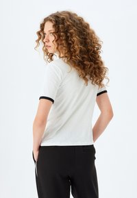 Polo de manga corta blanca con puños negros acanalados, que presenta una textura suave y un ajuste clásico. La parte trasera muestra un diseño simple y limpio.