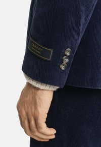 Ärmel eines marineblauen Cordjackets mit einem GANT-Label, ausgestattet mit drei abgerundeten Knöpfen und einem cremefarbenen Wollbund darunter für einen Texturkontrast.