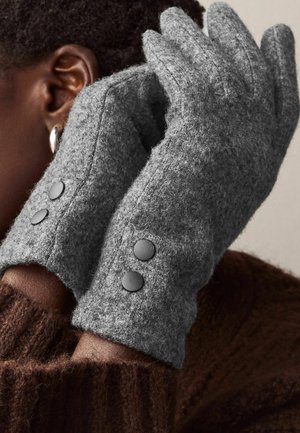Grau Wolle Handschuhe mit einer weichen Textur haben an jedem Manschetten zwei schwarze Knöpfe. Tragen von einer Person mit braunem Strickstoff im Hintergrund.