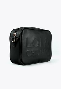Crna pravokutna kožna torbica s utisnutim slovima "LOLA CSDMNT", s patentnim zatvaračem na vrhu i malim metalnim prstenom sa strane.