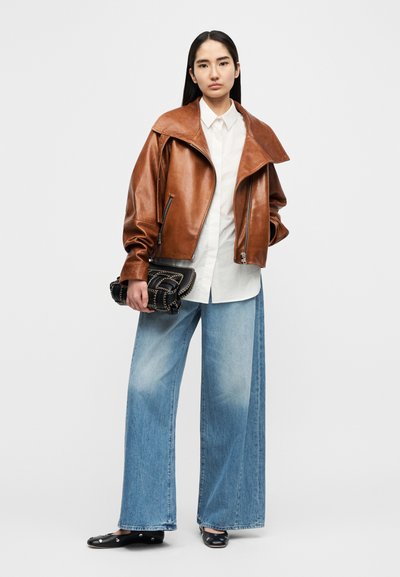 Καφέ δερμάτινο μπουφάν με υπερμεγέθη γιακά, λευκό πουκάμισο, φαρδιά ανοιχτό μπλε τζιν παντελόνια και μαύρο τσαντάκι χειρός, σε απλό φόντο.