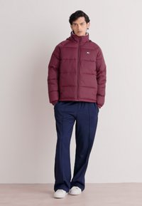 Bordeauxfarbene Steppjacke mit horizontalem Quilt-Design, Reißverschluss vorne und Kapuze. Kombiniert mit navyfarbener Hose und weißen Sneakern. Schlichtes, modernes Aussehen.