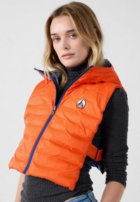 Chaleco acolchado naranja con cremallera azul y capucha, con un logo en el pecho izquierdo y textura acolchada. Se lleva sobre un jersey de cuello alto oscuro.