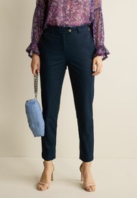 Pantaloni tapered blu navy con chiusura a bottone, abbinati a una blouse floreale trasparente e una borsa azzurra chiara con manico a catena.