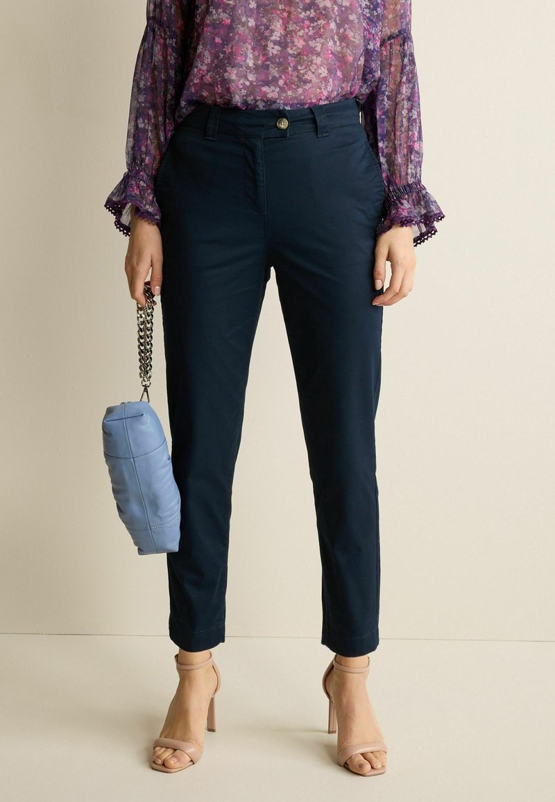 Pantaloni tapered blu navy con chiusura a bottone, abbinati a una blouse floreale trasparente e una borsa azzurra chiara con manico a catena.