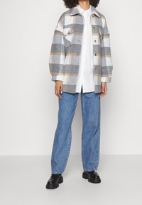 Chaqueta a cuadros en tonos grises, blancos y naranjas, con un cuello clásico, dos bolsillos en el pecho y botones. Combinada con jeans azules y botas negras.
