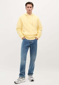 Gele hoodie met een voorzak, gecombineerd met lichtblauwe jeans. De outfit wordt aangevuld met grijze en witte sneakers.