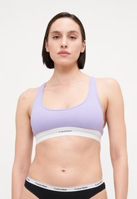 Fialová rebrová športová podprsenka so širokým bielym pásom s logom "Calvin Klein". Čierne spodné prádlo s logom na páse viditeľné dole.