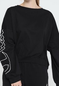 Schwarzes, kurz geschnittenes Sweatshirt mit langen Ärmeln, das ein weißes, abstraktes Design am linken Ärmel aufweist. Hergestellt aus weichem, strukturiertem Stoff.