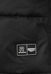 Detalje af en sort pufferjakke med et rektangulært stofmærke, der har "R PERFORMANCE" og "RESPECT" i hvid tekst, hvilket viser et minimalistisk design.