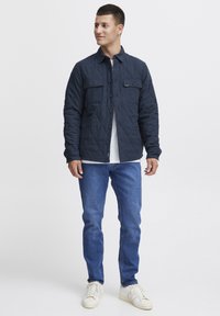 Quilted navy jacket with two chest pockets, button closure, worn over a white t-shirt, paired with blue jeans and cream sneakers.