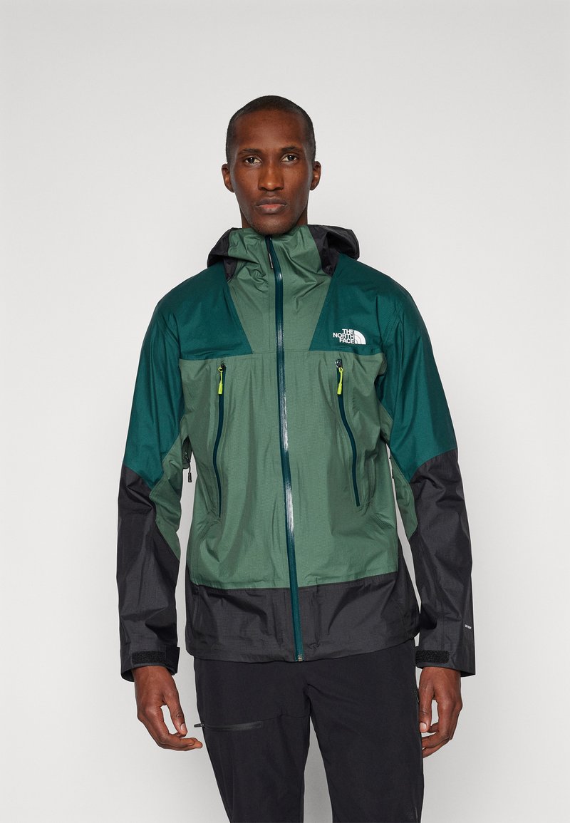 Veste imperméable verte et noire avec fermeture éclair à l'avant, deux poches poitrine et capuche réglable. Fabriquée en tissu respirant avec des accents contrastés.