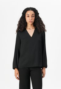 Jonge vrouw met lang krullend haar, gekleed in een zwarte V-hals lange mouw blouse en zwarte broek, staand tegen een witte achtergrond.