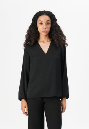 Young woman with long curly hair wearing a black V-neck long-sleeve blouse and black pants, standing against a white background.