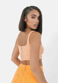 Pfirsichfarbener Cropped Top mit Reißverschluss auf der Rückseite, geripptem Stoff und dünnen Trägern, kombiniert mit hochgarzigen orangefarbenen Jeans. Glatte Textur und taillierte Passform.