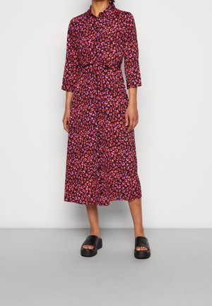 Vrouw draagt een midi-jurk met rode en paarse luipaardprint, 3/4 mouwen en zwarte platform slides, staand tegen een lichte achtergrond.