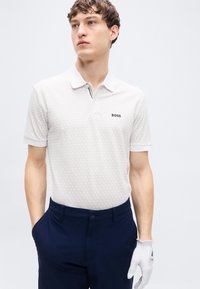 Πολό κοντομάνικο σε ανοιχτό γκρι με διακριτικό εξαγωνικό μοτίβο, με λογότυπο της μάρκας και κολάρο με ριγέ αντίθεση. Συνδυασμένο με ναυτικό μπλε παντελόνι και γάντι.