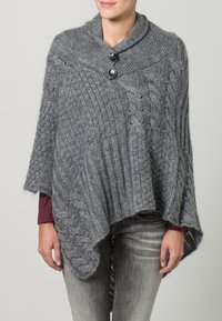 Grå stickad poncho med sjalkrage, två svarta knappar, kabelmönster och asymmetrisk fåll. Texturerat tyg med ribbmönster.