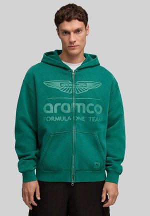 Młody mężczyzna w zielonym kapturze zapinanym na zamek z logo zespołu Aston Martin Aramco Formula One, stoi na jednolitym szarym tle.