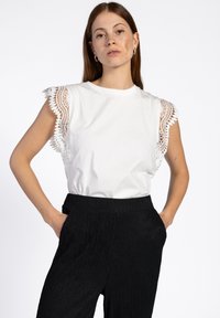 Witte katoenen t-shirt met kantdetails op de schoudernaden, gecombineerd met zwarte ribgebreide wijde broek, weergegeven tegen een effen achtergrond.