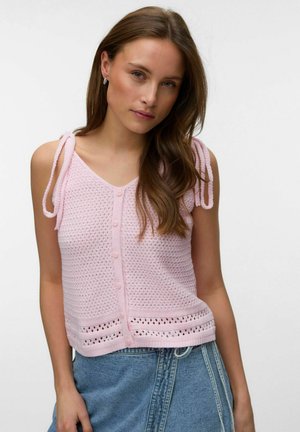 Młoda kobieta ubrana w różowy, bezrękawnik dzianinowy z guzikami i wiązanymi ramiączkami, zestawiony z wysokim stanem niebieskich dżinsów.