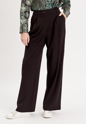 Persoon draagt zwarte wijde broek met voorplooien, één hand in de zak, gecombineerd met witte sneakers en een groen top met patroon.