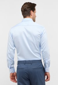 Camicia azzurra chiara con maniche lunghe, polsini con bottone e una texture liscia. Presenta un collo classico e un dettaglio discreto nella parte posteriore.