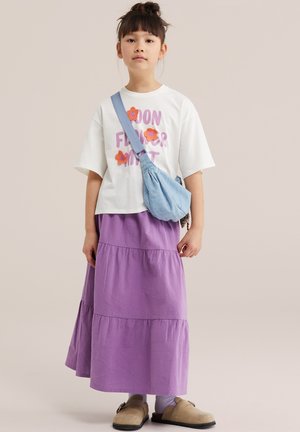 Mladé dievča v bielom tričku s kvetinovým vzorom, fialovej viaczvrstvovej sukni, modrej kabelke cez rameno a béžových nazúvacích topánkach stojí na neutrálom pozadí.