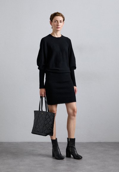 Donna che indossa un maglione nero, una gonna a coste, stivali alla caviglia, mentre tiene una borsa tote nera trapuntata, in piedi davanti a uno sfondo grigio chiaro uniforme.