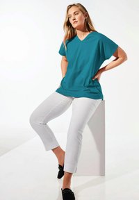 Türkises V-Ausschnitt T-Shirt aus weichem Material, kombiniert mit weißen, knöchelfreien Hosen. Schwarze Slipper mit dekorativen Akzenten runden das Outfit ab.
