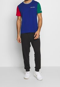 Muž na sobě modré tričko Champion s červenými a zelenými rukávy, černé jogger kalhoty Adidas a bílé tenisky, stojí před jednobarevným pozadím.