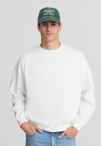 Felpa bianca oversize con scollo tondo e maniche a sbuffo, abbinata a un berretto verde scuro con testo bianco. Jeans in denim.